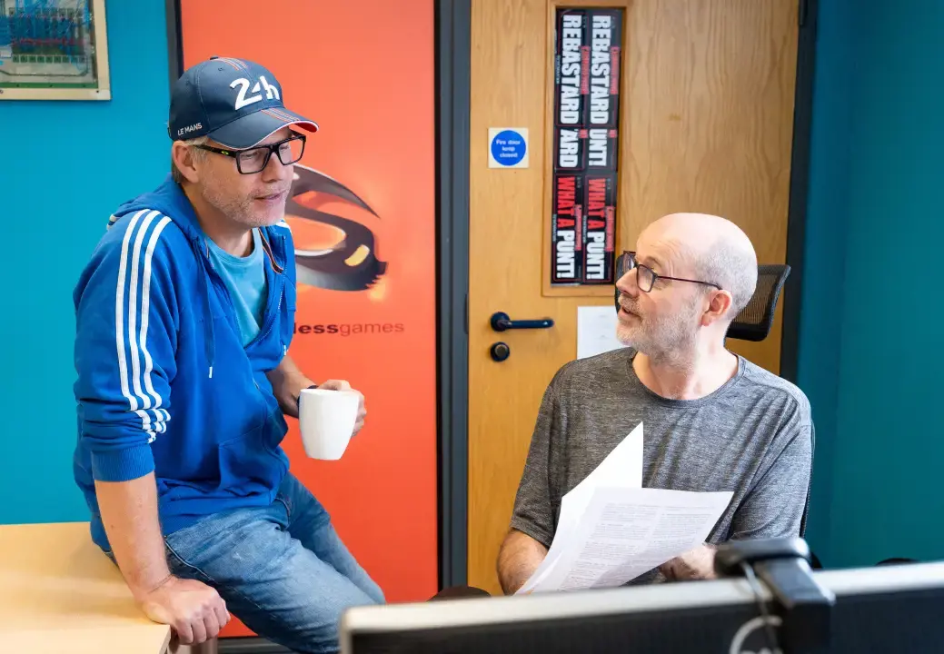 Photo of two Stainless Games team members talking to each other. One wearing a cap sat on a desk holding a mug. The other sat on a chair holding papers that they are both looking at.